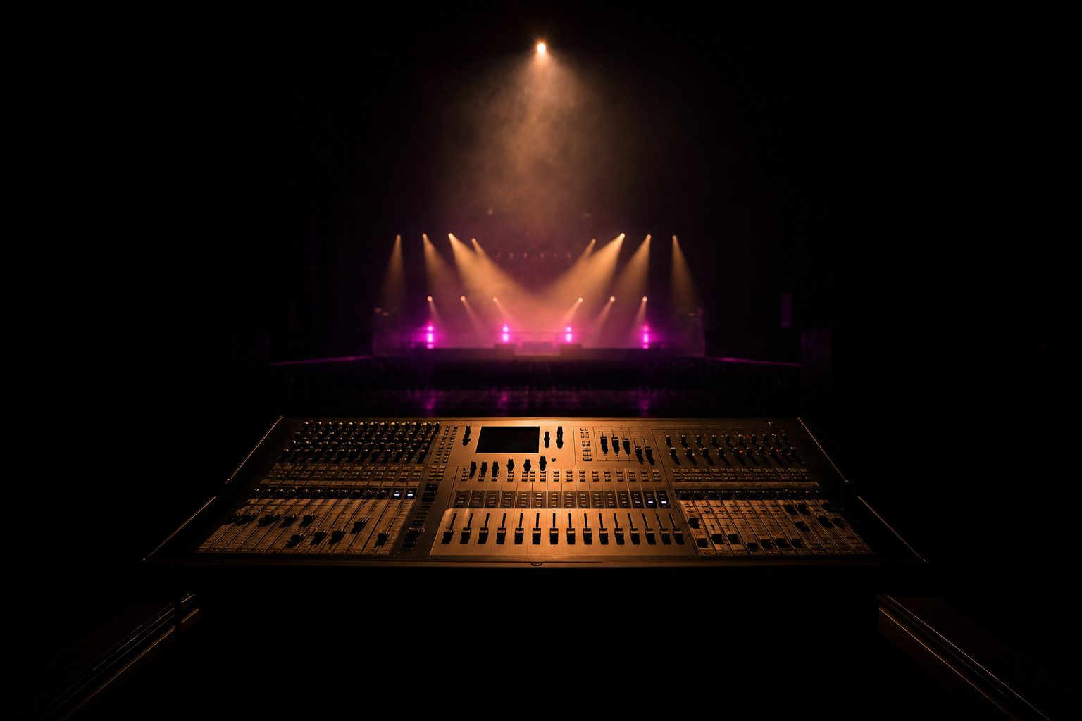 An empty front-of-house desk silhouetted against a distant lit stage, its faders catching a sliver of warm light.