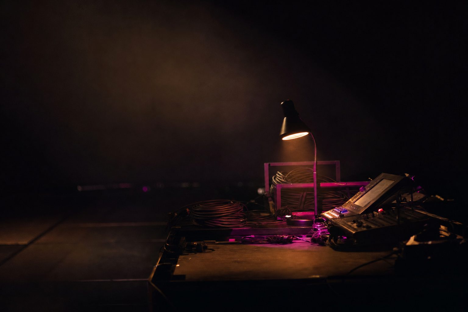 A lone desk lamp glow on a darkened production desk, cables coiled, everything else in black.