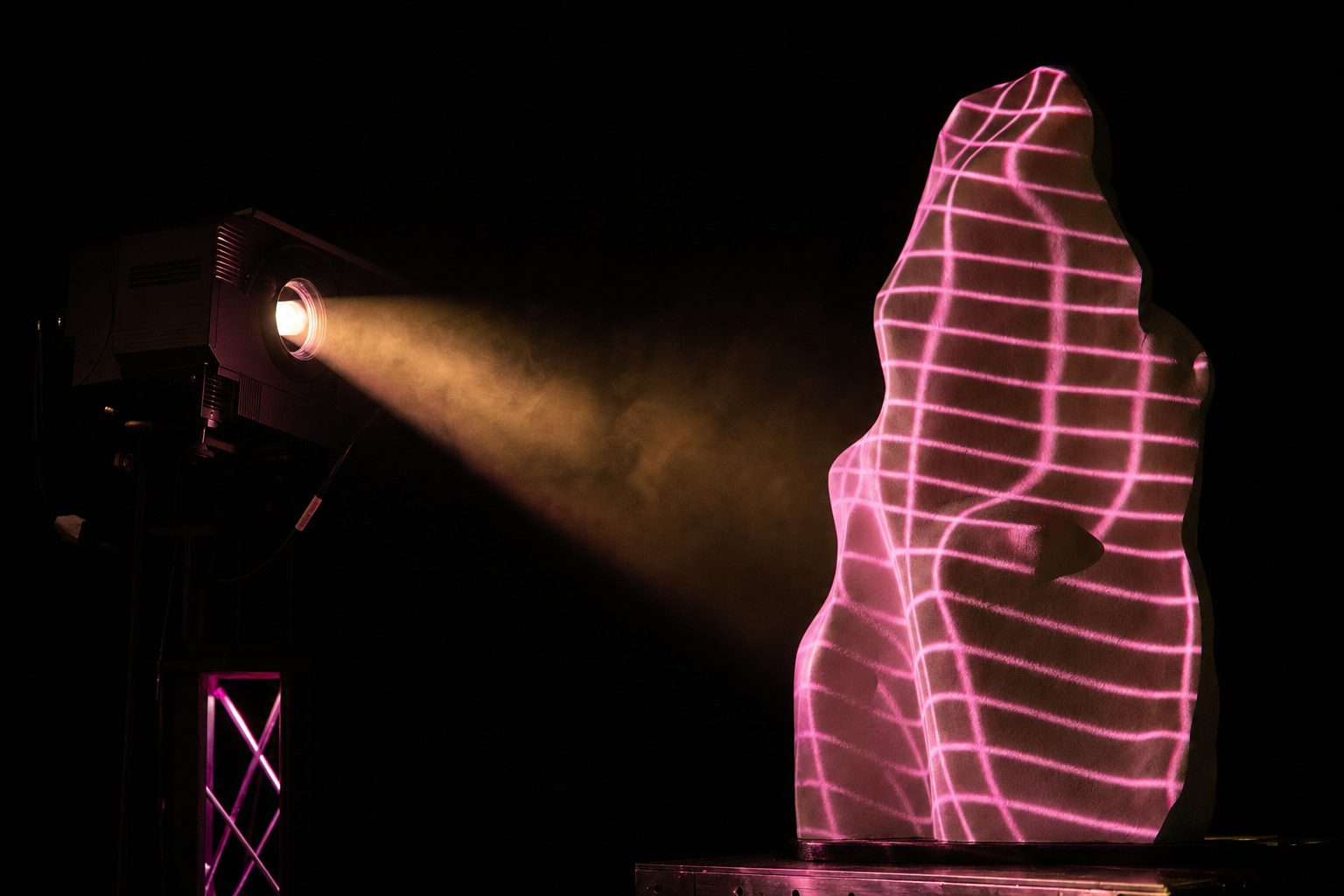 A projector throwing a warped grid of light onto an irregular white sculptural object in a dark room.