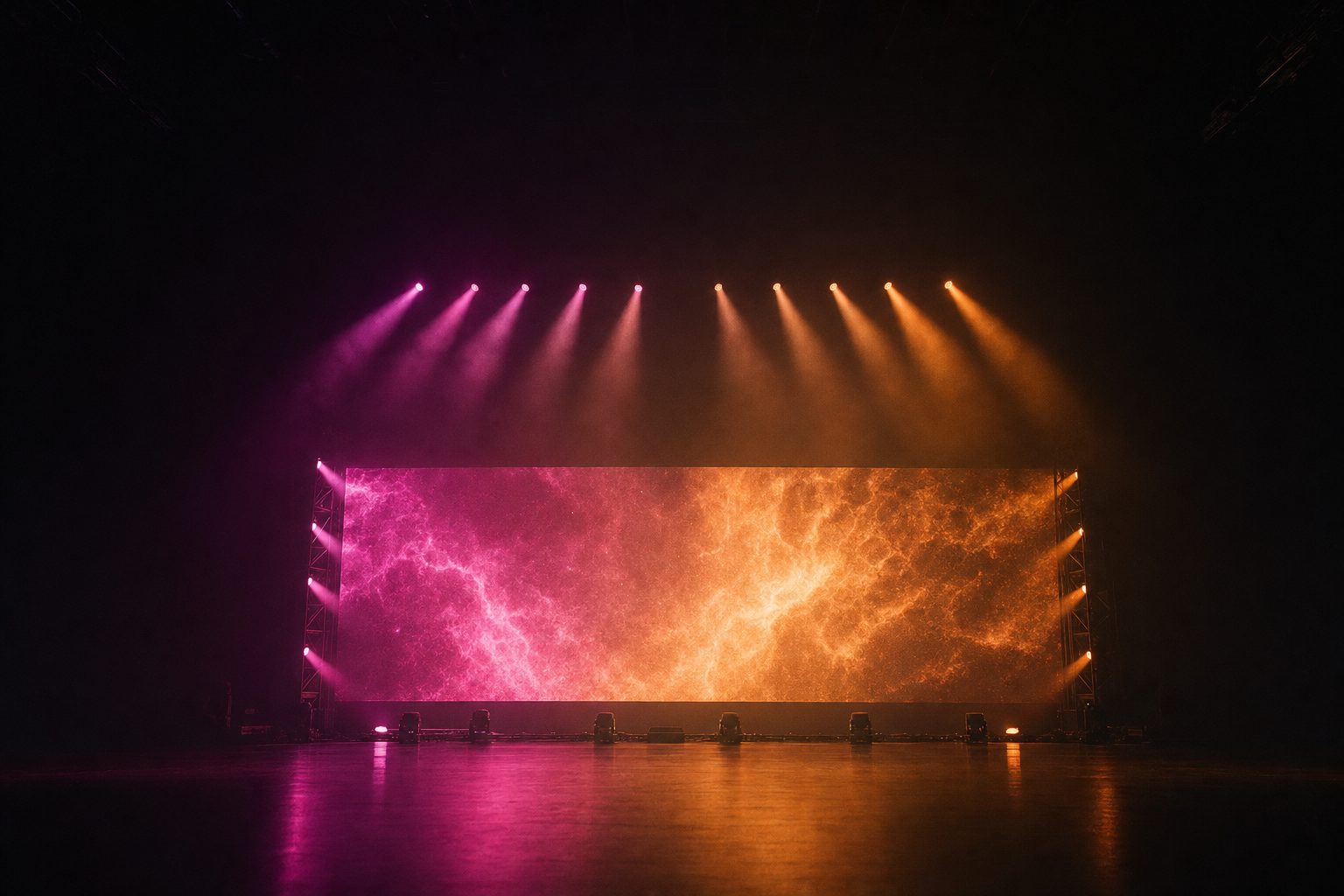 A wide LED wall seen head-on across an empty dark arena floor, with magenta and amber beams fanning down through heavy haze.