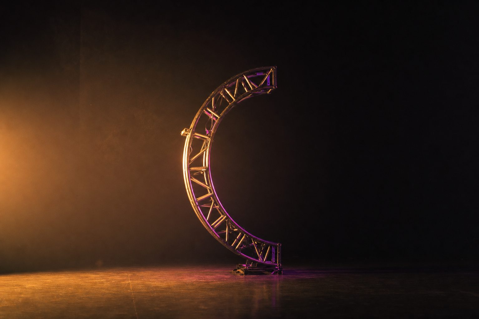 A single large sculptural object standing alone on a bare stage deck in an empty venue, lit from one low side so half of it is bright and half is lost, no rigging visible above it.