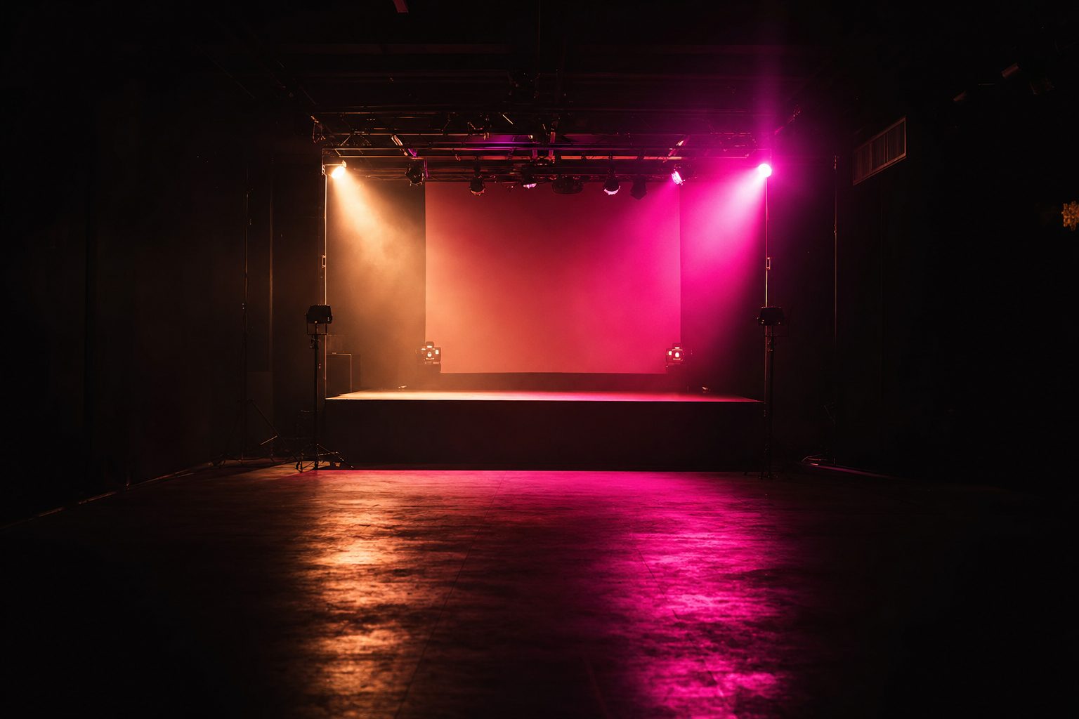 One small room built for a single night: a low stage, a modest lighting position and a single screen surface, seen from the back of an empty floor with the house lights off.