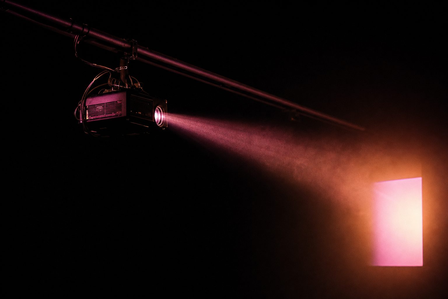 A single projector on a rigging bar in a dark room, its beam widening across a measured distance toward a bright rectangle of light on a far surface.