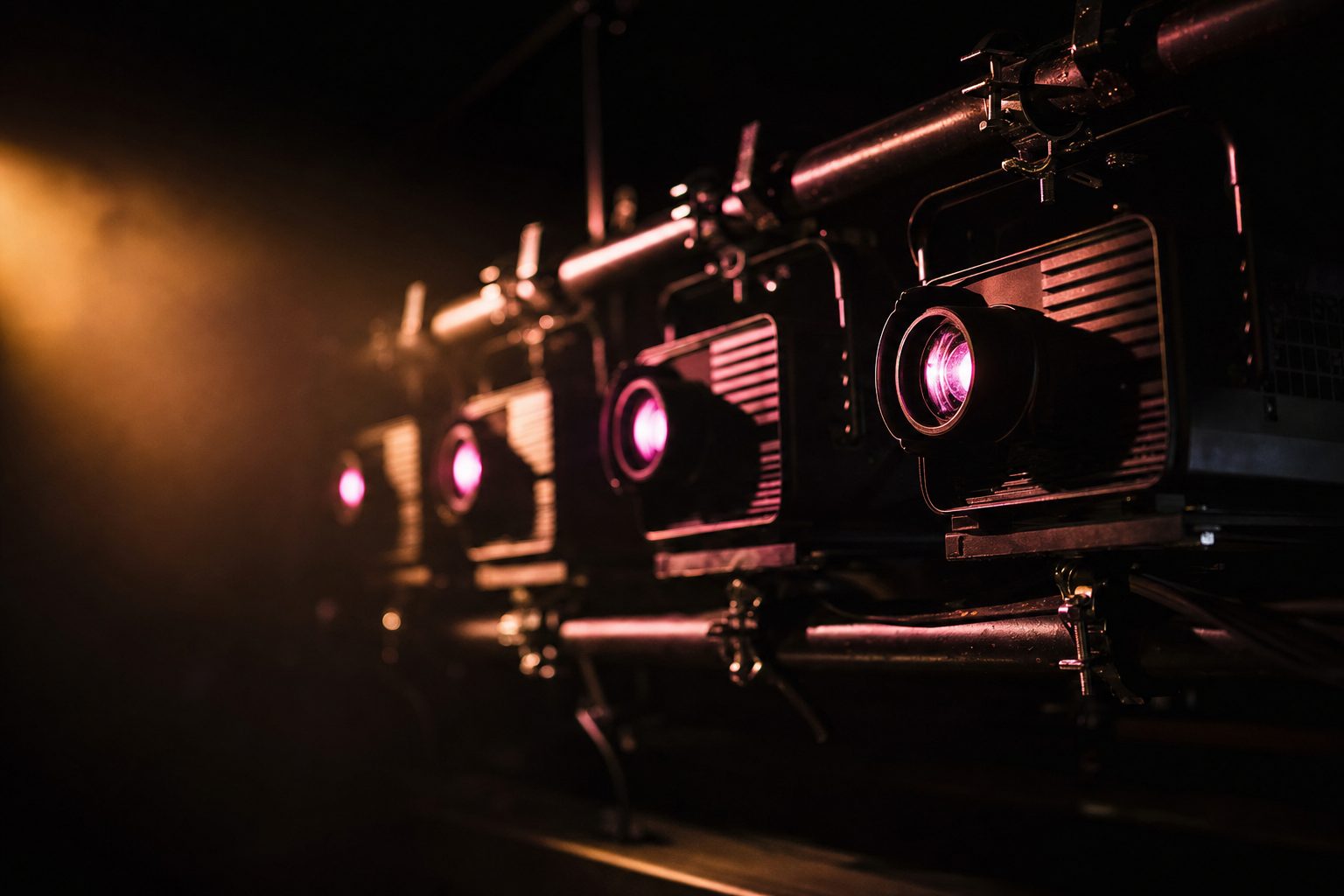 A row of dormant projectors on a rigging bar, their lenses catching a single magenta highlight.